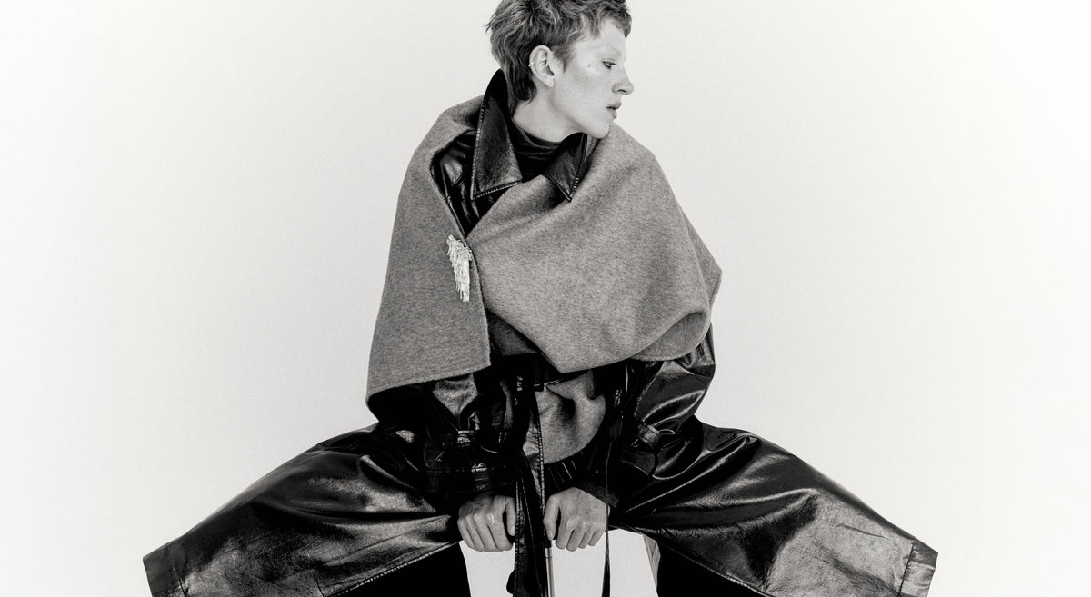 A black and white editorial fashion photograph of a model wearing a voluminous, wide-leg black leather outfit with a large draped gray woolen shawl or cape, seated against a plain light background.