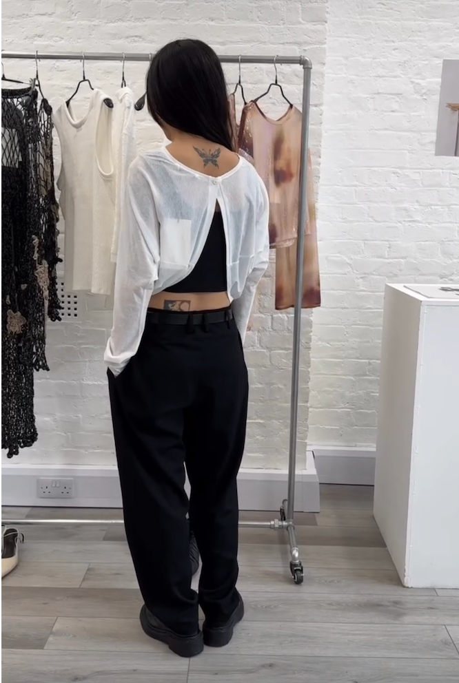 A woman stands with her back to the camera wearing a sheer white long-sleeve crop top over a black crop top and black wide-leg trousers, standing in front of a clothing rack in a bright room with a white brick wall.