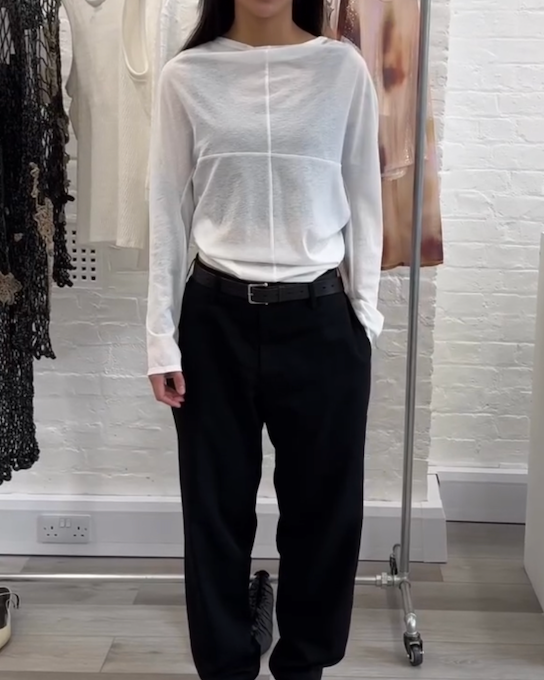 A person standing in a clothing store wearing a white long-sleeve top, black wide-leg pants, and black shoes.