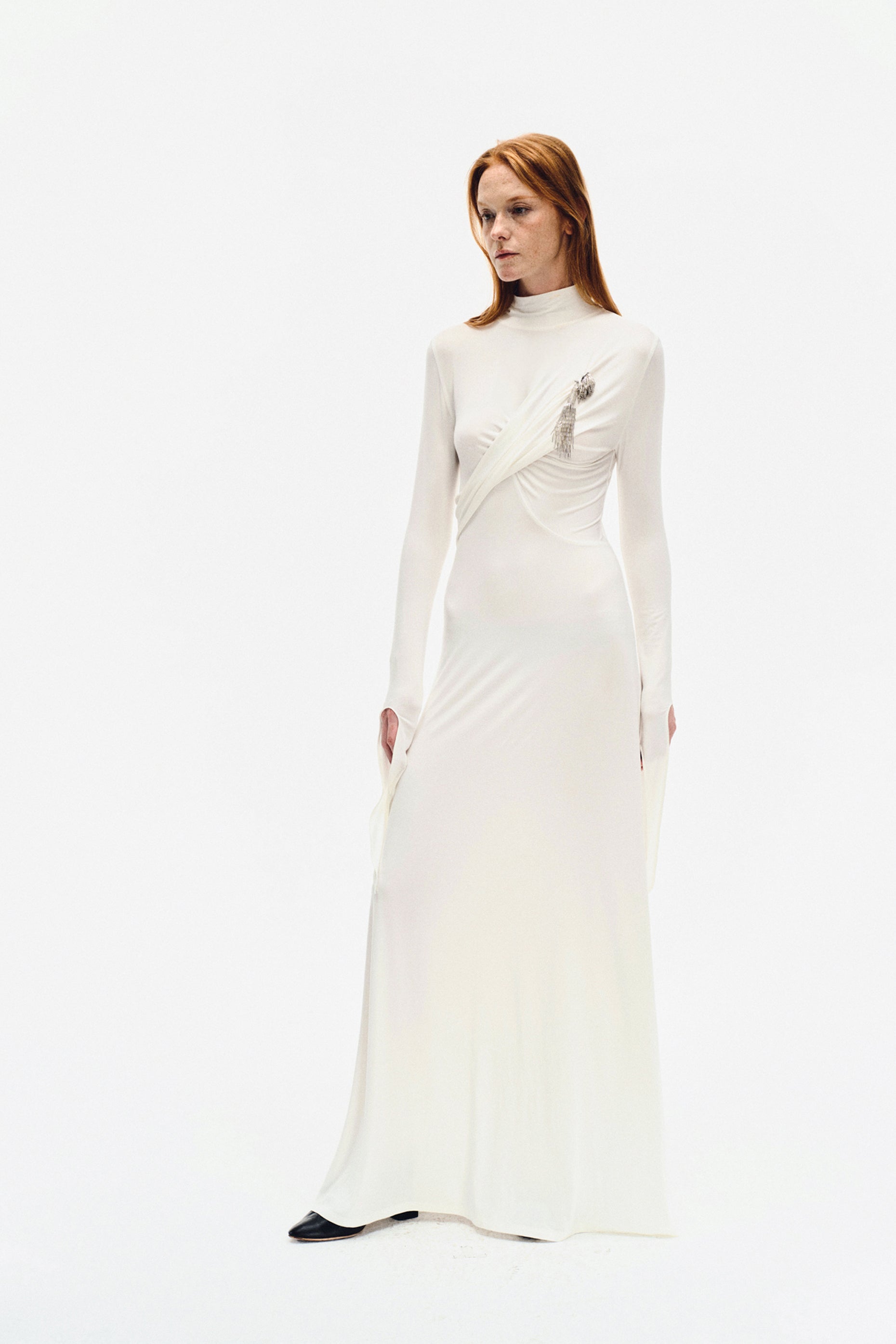 Side angle of YIBRI ivory long-sleeve maxi dress featuring soft draping, high neckline and sculptural wrap detail with silver brooch ornament