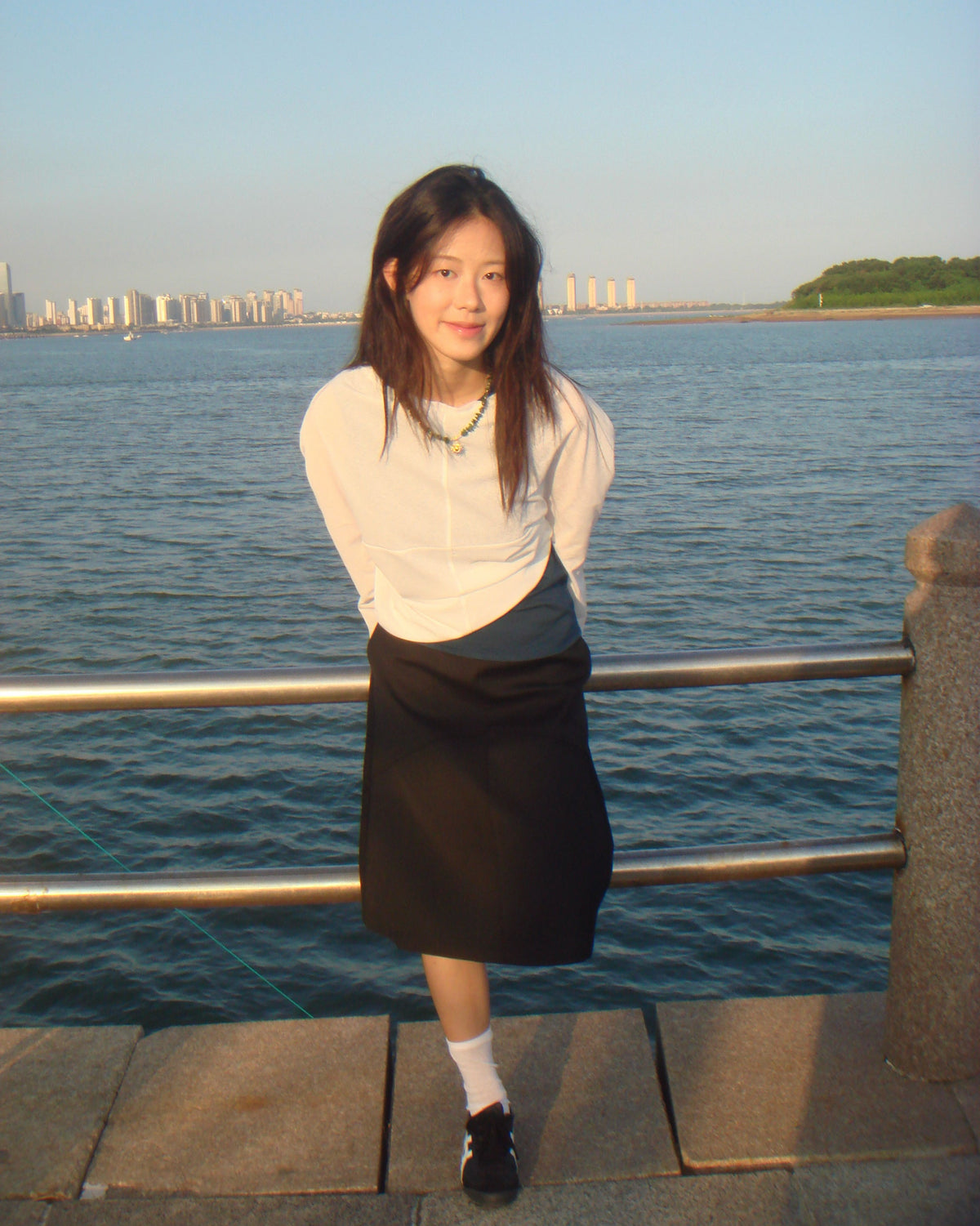 A woman stands outdoors by a waterfront railing, wearing a white long-sleeve top and a black skirt. The background shows water, distant buildings, and a clear sky.