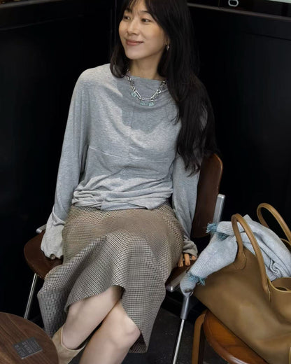 A woman sits on a wooden chair wearing a light gray long-sleeve knit top and a brown houndstooth skirt. She is accessorized with a silver geometric necklace and beige ankle boots.