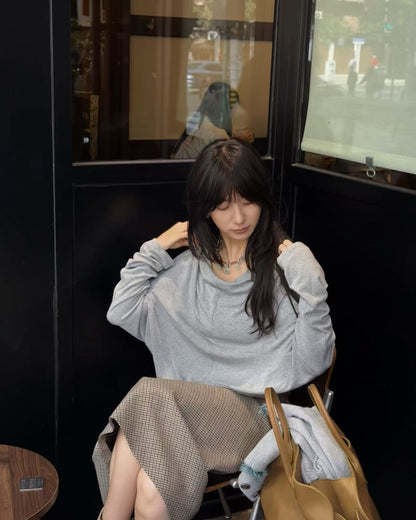 A woman sitting in a cafe setting wearing a light gray oversized long-sleeve top and a brown houndstooth skirt, with a tan leather tote bag next to her.