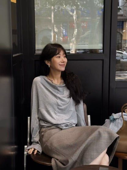 A woman sitting in a chair in front of a dark window, wearing a light gray draped long-sleeve top and a brown and beige checkered midi skirt.