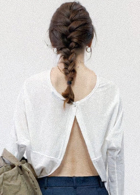 A woman shown from behind wearing a white long-sleeve cropped top with a large open back, paired with dark blue jeans. Her hair is styled in a braided ponytail.