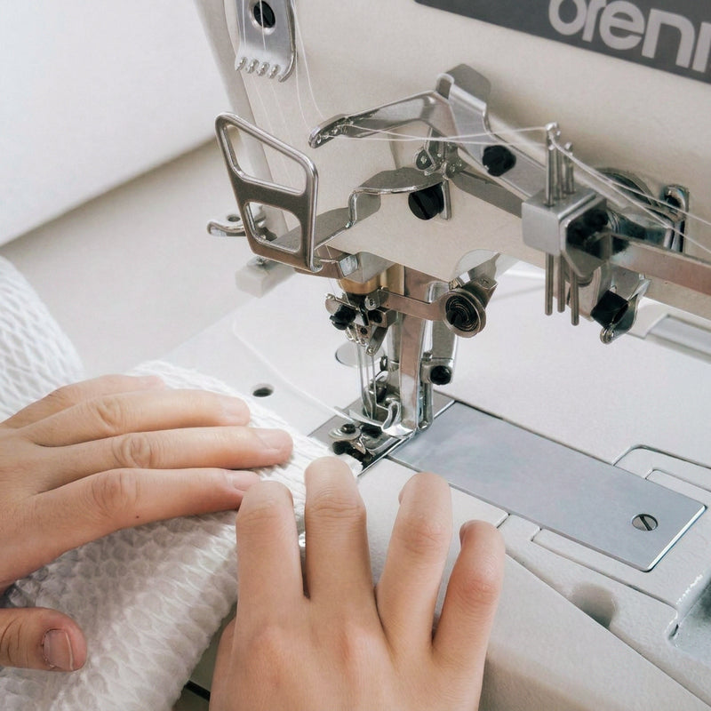 Close-up of a person's hands guiding white textured fabric under a silver and white sewing machine with metallic presser feet.